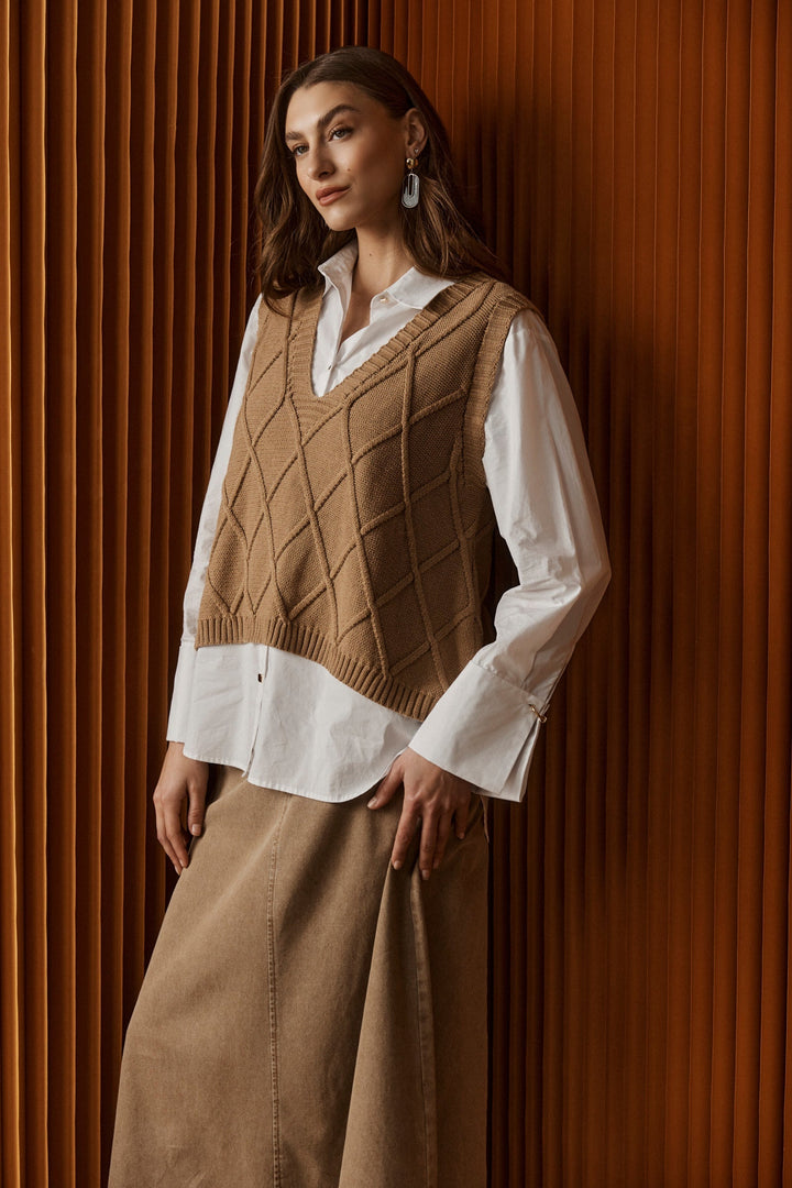 Model wearing a women's mocha V-neck knit vest layered over a white shirt against a textured backdrop
