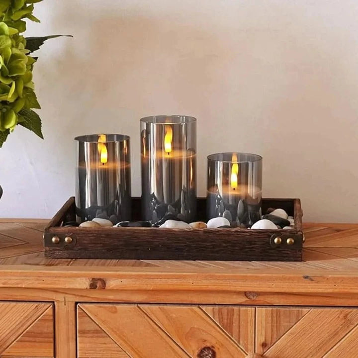Three gray glass LED flameless candles arranged on a wooden tray with decorative stones.