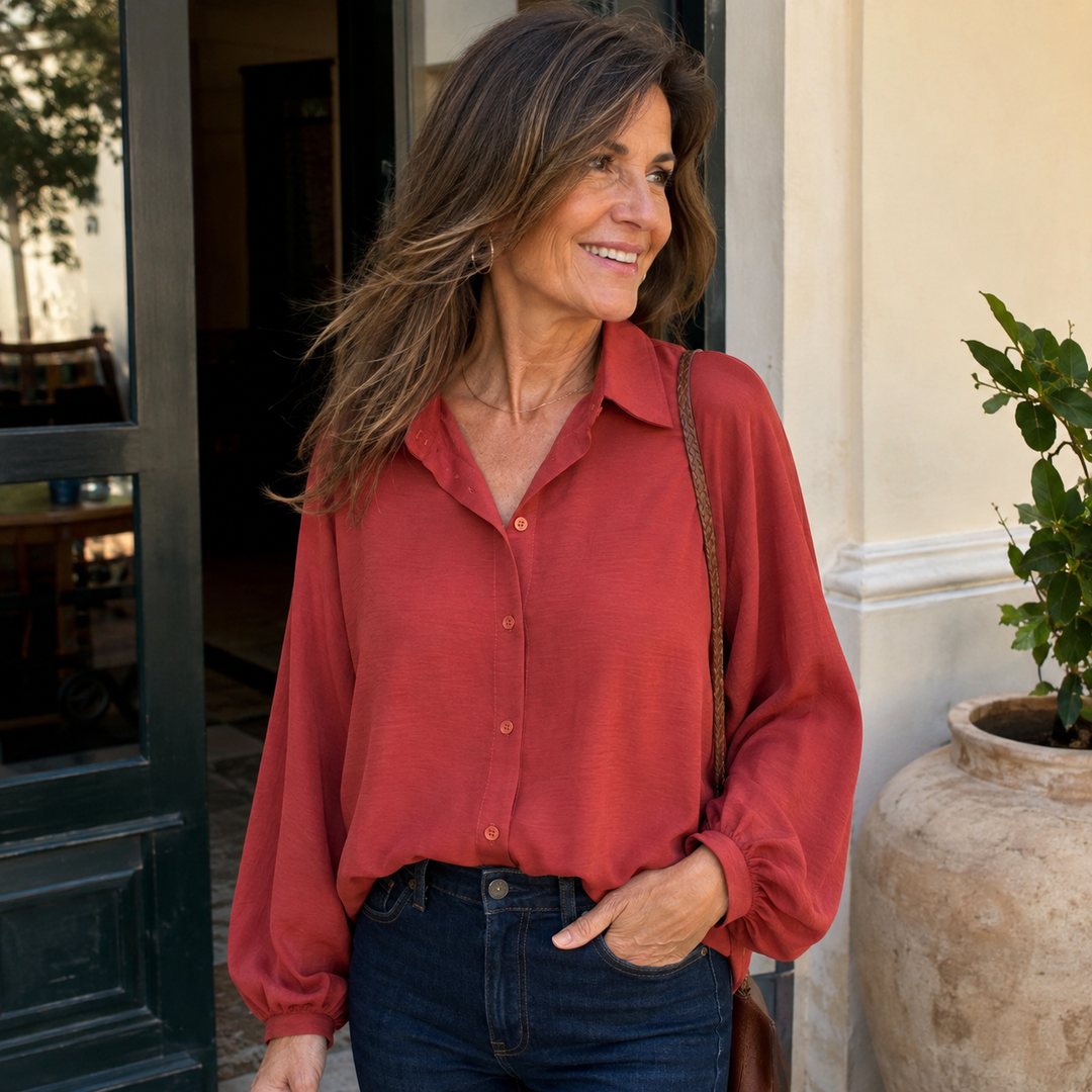 A smiling woman wearing a red long sleeve button down shirt paired with dark jeans outdoors.