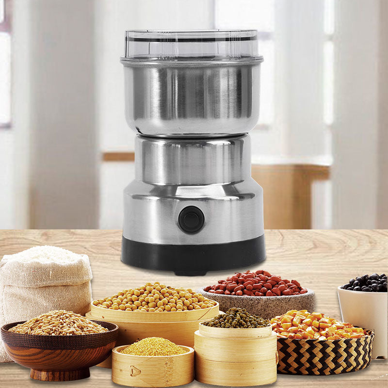 Portable electric kitchen grinder in silver finish surrounded by raw grains and seeds on a wooden table.