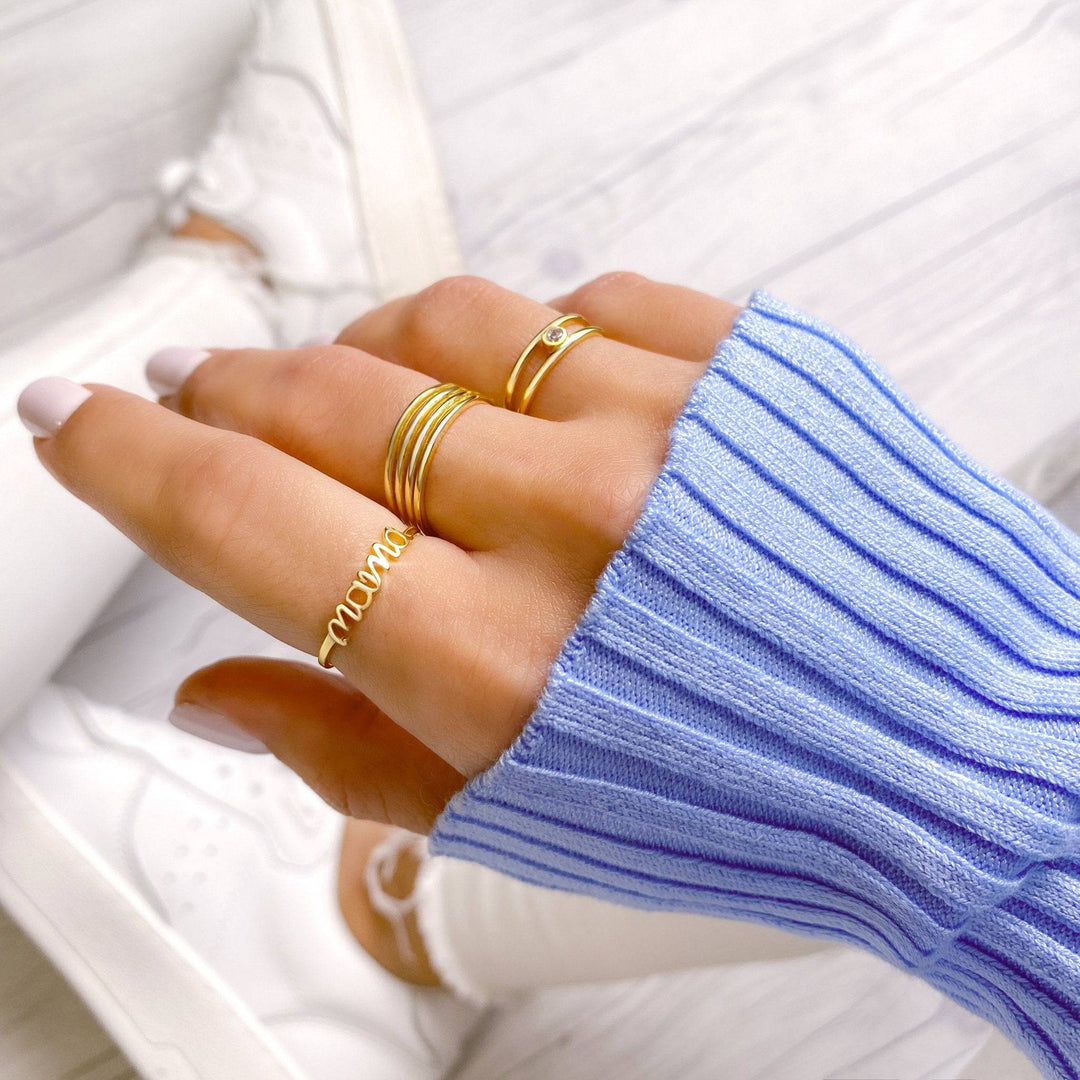A woman's hand wearing a gold Mama ring alongside other delicate gold bands and a blue sweater.