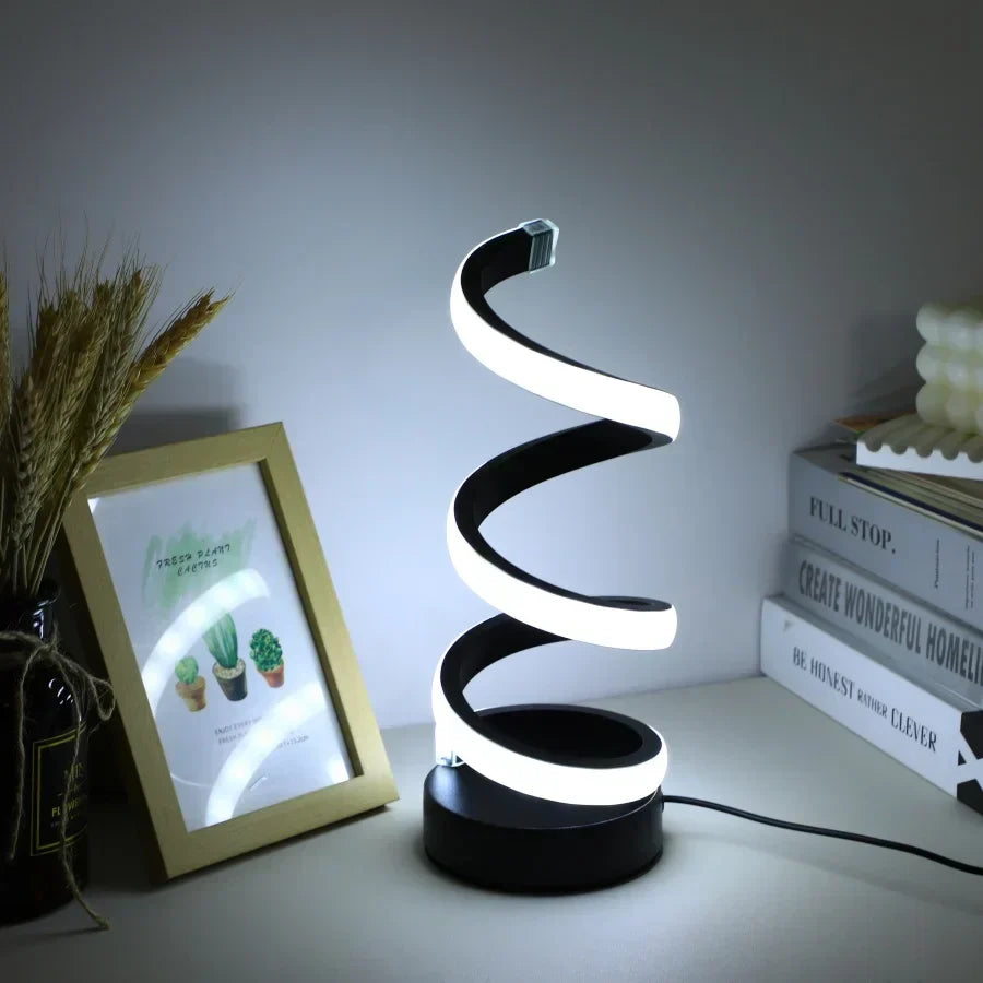 Modern S-shaped black desk lamp glowing on a wooden desk next to picture frames and books