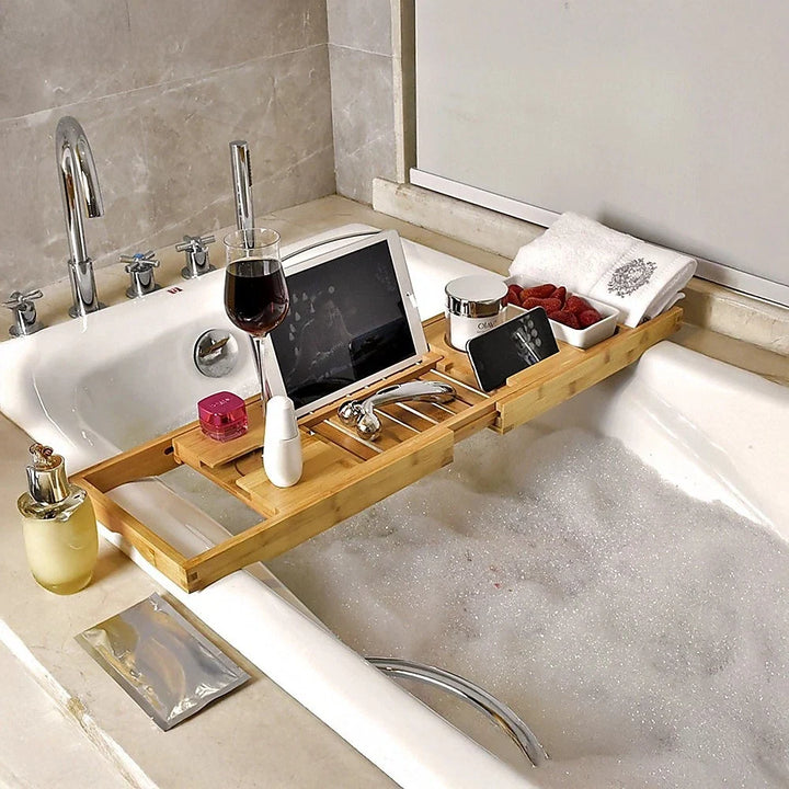 Expandable bamboo bathtub caddy holding a tablet, wine glass, and cosmetics over a full tub