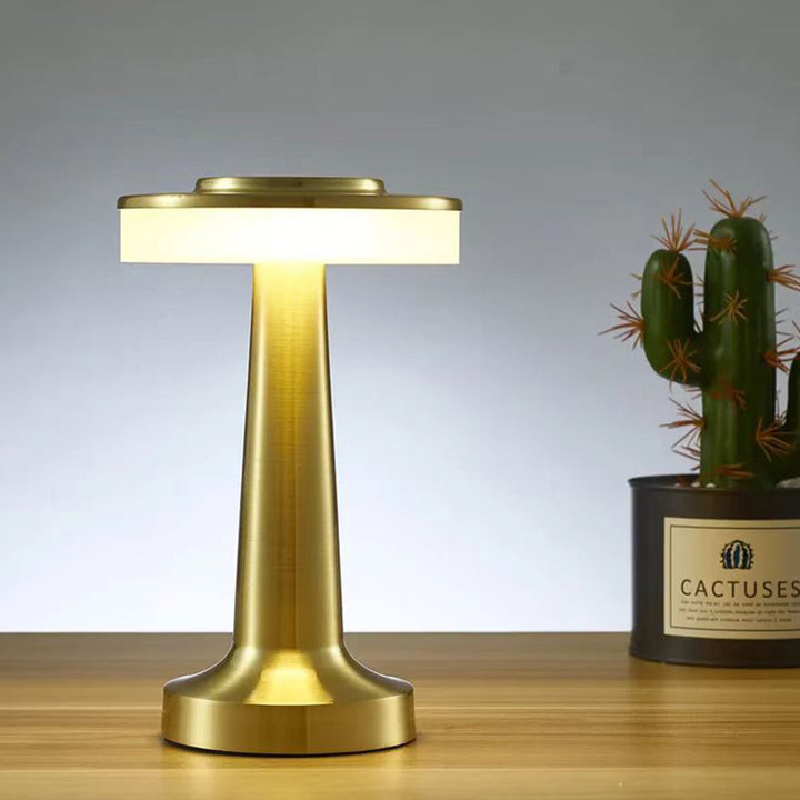 Gold rechargeable touch LED table lamp standing on a wooden surface with a potted cactus in the background.