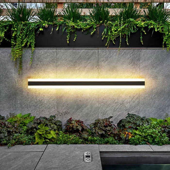 Extra long vertical outdoor wall lamp mounted on a stone wall above lush greenery