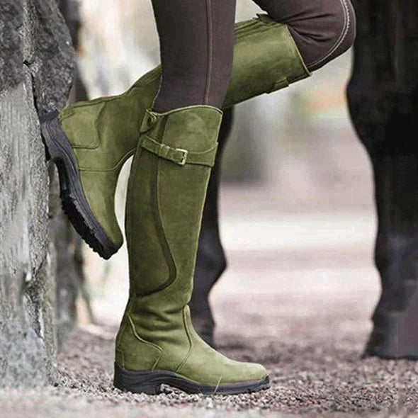 Woman wearing green waterproof tall riding boots outdoors against a stone wall
