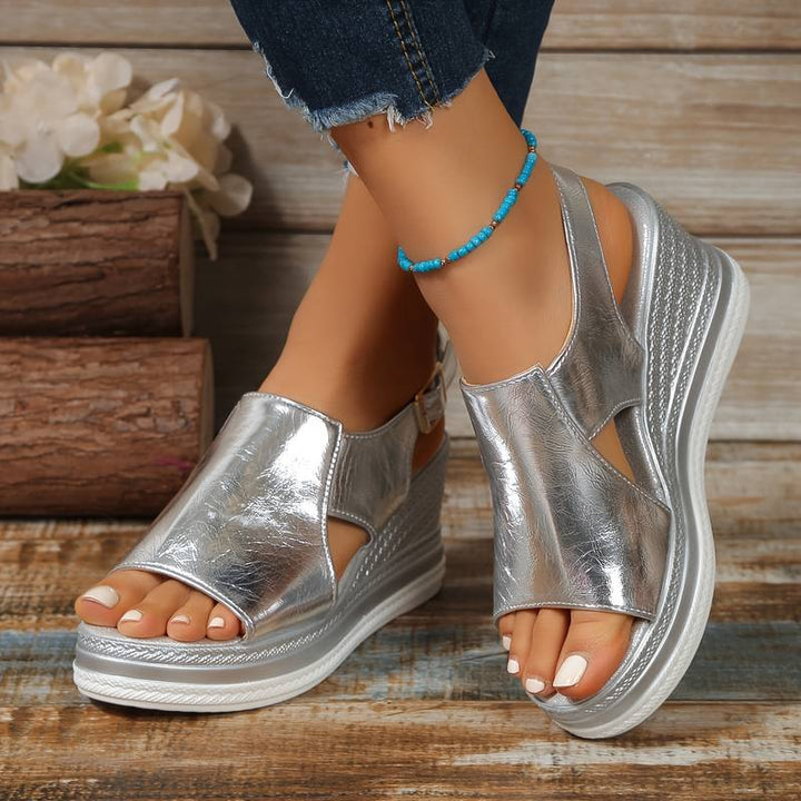 Model wearing shiny silver wedge platform sandals with cutout details and ankle straps