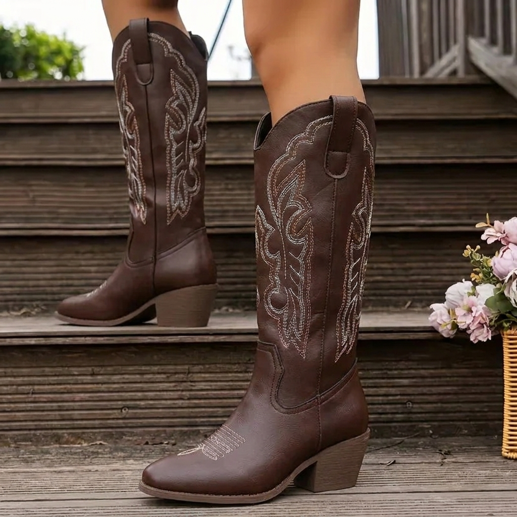 Brown women's knee-high cowboy boots with detailed scrollwork embroidery on wooden stairs