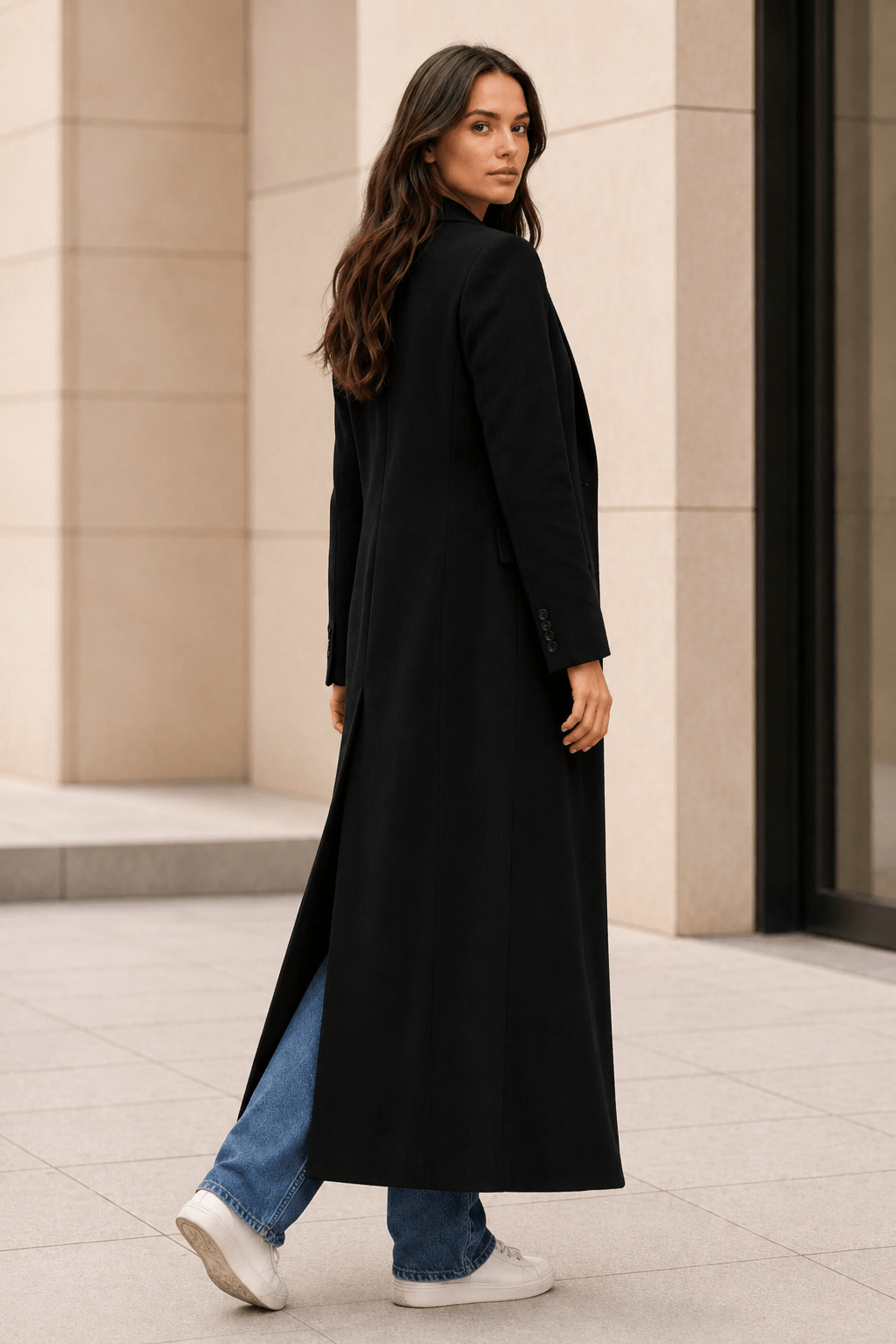 Full length rear view of a woman walking in a minimalist black long winter coat with structured shoulders.