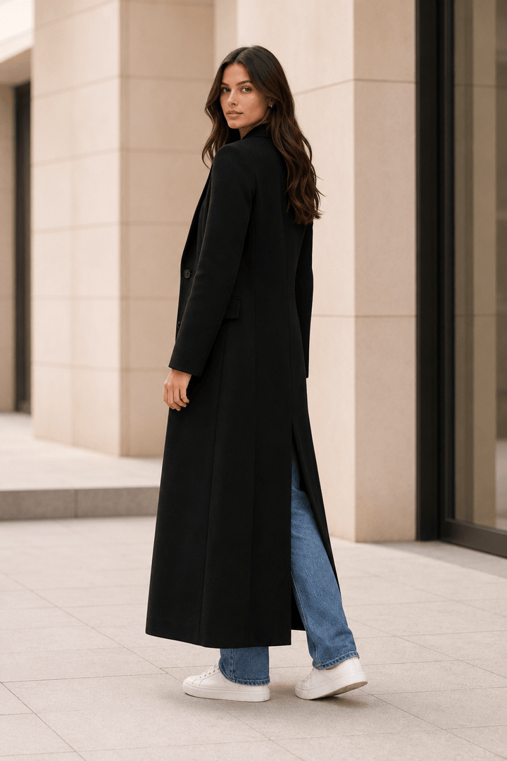 Side back view of a woman showcasing the floor-length drape and back vent of the black maxi coat.